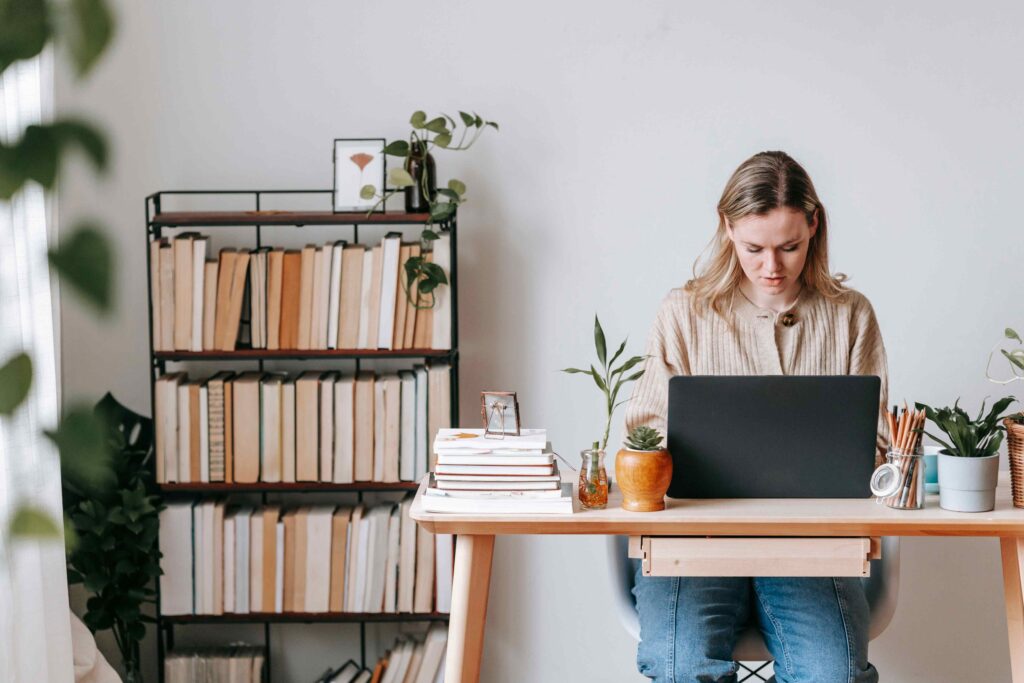 A woman concentrates on a laptop at a table in a home office with plants and a bookshelf behind her