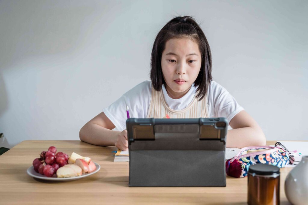 A young girl studies at a table while looking at a tablet