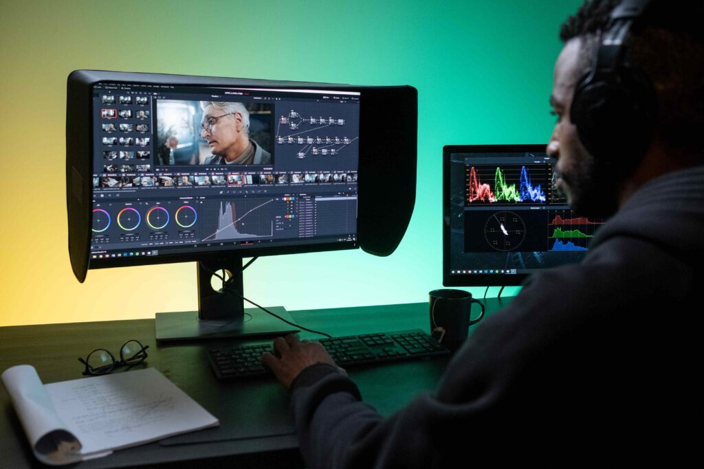 A man sits at a desk editing video footage across two monitors in a modern studio